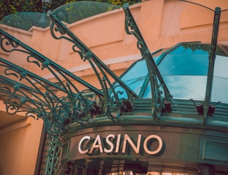The image displays the entrance to a casino, characterized by detailed and ornate architectural elements with green metal structures and a sign that reads 'CASINO'. The background includes a glass dome and a peach-colored wall.