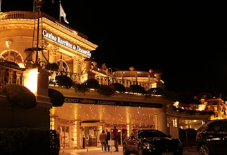 A luxurious casino at night, illuminated with warm, golden lights. The building features elegant architecture with columns and decorative elements. A few people are walking towards the entrance, and there are a couple of parked cars nearby.