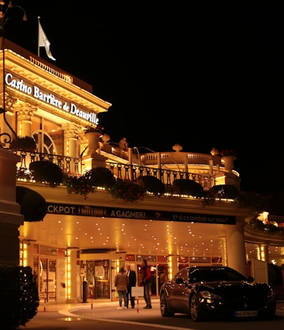 A luxurious casino at night, illuminated with warm, golden lights. The building features elegant architecture with columns and decorative elements. A few people are walking towards the entrance, and there are a couple of parked cars nearby.