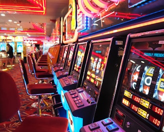 A brightly lit casino interior featuring a row of colorful slot machines. The neon lights create a vibrant atmosphere, with patterned carpeting adding to the decor. Stools are aligned neatly in front of each machine.