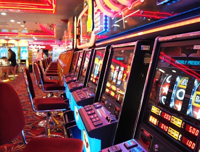 A brightly lit casino interior featuring a row of colorful slot machines. The neon lights create a vibrant atmosphere, with patterned carpeting adding to the decor. Stools are aligned neatly in front of each machine.