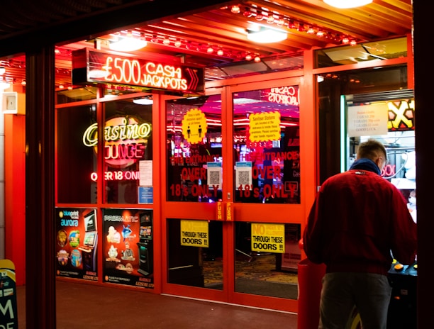 A brightly lit entrance to a casino with neon signs, including one advertising a £500 jackpot. The doors have clear signage indicating entry is restricted to those over 18. A person in a red jacket stands to the right, possibly interacting with a machine or display.