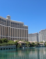 A large luxury hotel and casino building with multiple stories, featuring a light facade with arched windows. The structure has a prominent central tower with a dome-like top. It is surrounded by landscaping with trees and a body of water in the foreground.