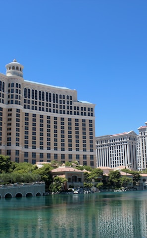 A large luxury hotel and casino building with multiple stories, featuring a light facade with arched windows. The structure has a prominent central tower with a dome-like top. It is surrounded by landscaping with trees and a body of water in the foreground.