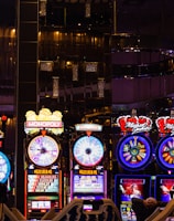 A lively casino scene featuring a row of bright, colorful slot machines with themes like Monopoly, Michael Jackson, and Lord of the Rings. The gaming machines display a variety of flashing lights and digital screens, creating an energetic and exciting atmosphere. Reflections of the lights are visible in glossy surfaces above the machines.