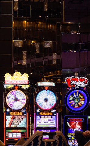 A lively casino scene featuring a row of bright, colorful slot machines with themes like Monopoly, Michael Jackson, and Lord of the Rings. The gaming machines display a variety of flashing lights and digital screens, creating an energetic and exciting atmosphere. Reflections of the lights are visible in glossy surfaces above the machines.