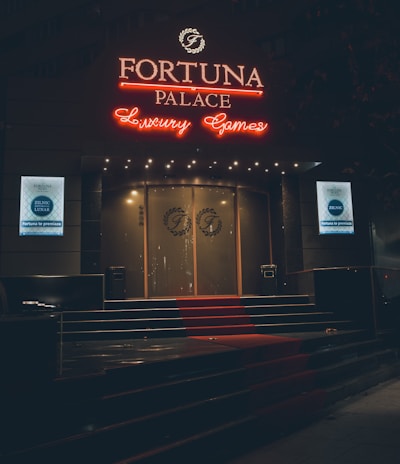 A neon-lit casino entrance with a red carpet leading up to large double doors. The sign reads 'Fortuna Palace Luxury Games' in bright red and white neon lights. The dimly lit atmosphere suggests a night-time setting. There are two digital displays on either side of the entrance showing promotional content.