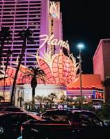 A brightly lit casino with neon lights in vibrant red, pink, and blue, featuring a large flamingo signage. Palm trees are visible in the foreground with vehicles passing by on the street at night.