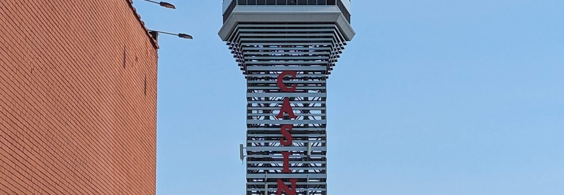 A tall tower with a modern design features the large vertical red letters spelling 'CASINO'. The structure has a sleek, metallic appearance with various horizontal and vertical lines. The sky is mostly clear with a few clouds. In the foreground, part of a brick building is visible, along with some trees and buildings in the distance.