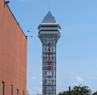 A tall tower with a modern design features the large vertical red letters spelling 'CASINO'. The structure has a sleek, metallic appearance with various horizontal and vertical lines. The sky is mostly clear with a few clouds. In the foreground, part of a brick building is visible, along with some trees and buildings in the distance.