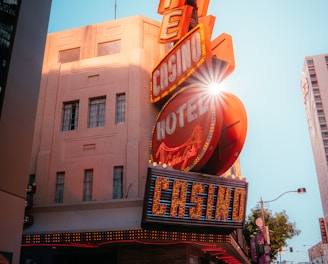A vibrant city scene featuring a tall, illuminated casino sign with retro-style neon lights. The sign dominates the building facade with the words 'CASINO' and 'HOTEL'. Sunlight peeks through, creating a dramatic lens flare effect. The street is lively, with pedestrians casually walking and a hint of urban hustle.