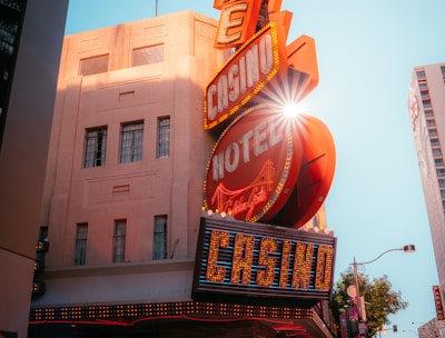 A vibrant city scene featuring a tall, illuminated casino sign with retro-style neon lights. The sign dominates the building facade with the words 'CASINO' and 'HOTEL'. Sunlight peeks through, creating a dramatic lens flare effect. The street is lively, with pedestrians casually walking and a hint of urban hustle.